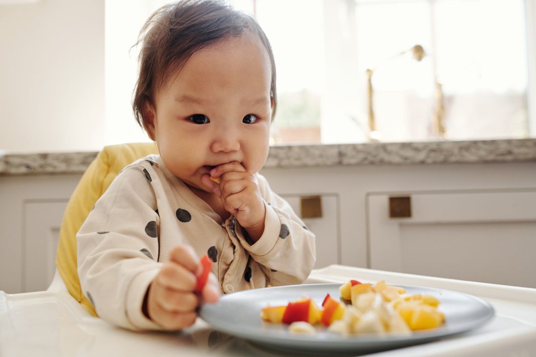 A baby in a polka-dot outfit eats fresh fruit in a high chair